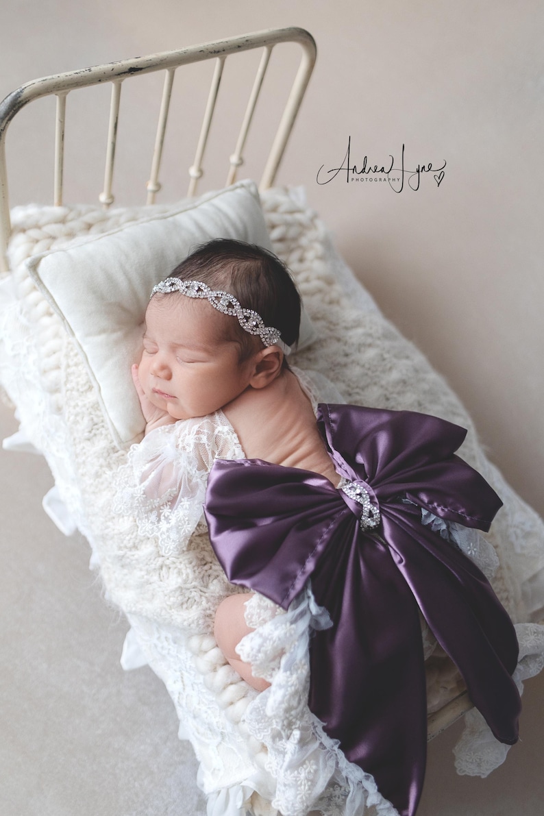 May include: A newborn infant wearing a white lace wrap with a large, deep purple satin bow on the back and a silver-toned rhinestone headband. The infant is resting on a cream-colored chunky knit blanket inside a small, vintage-style metal bed frame.