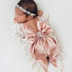 May include: A cream-colored lace romper featuring long sleeves with ruffled edges and a large, dusty rose satin bow on the back. The outfit is accented with a central rhinestone embellishment on the bow and a matching rhinestone headband.