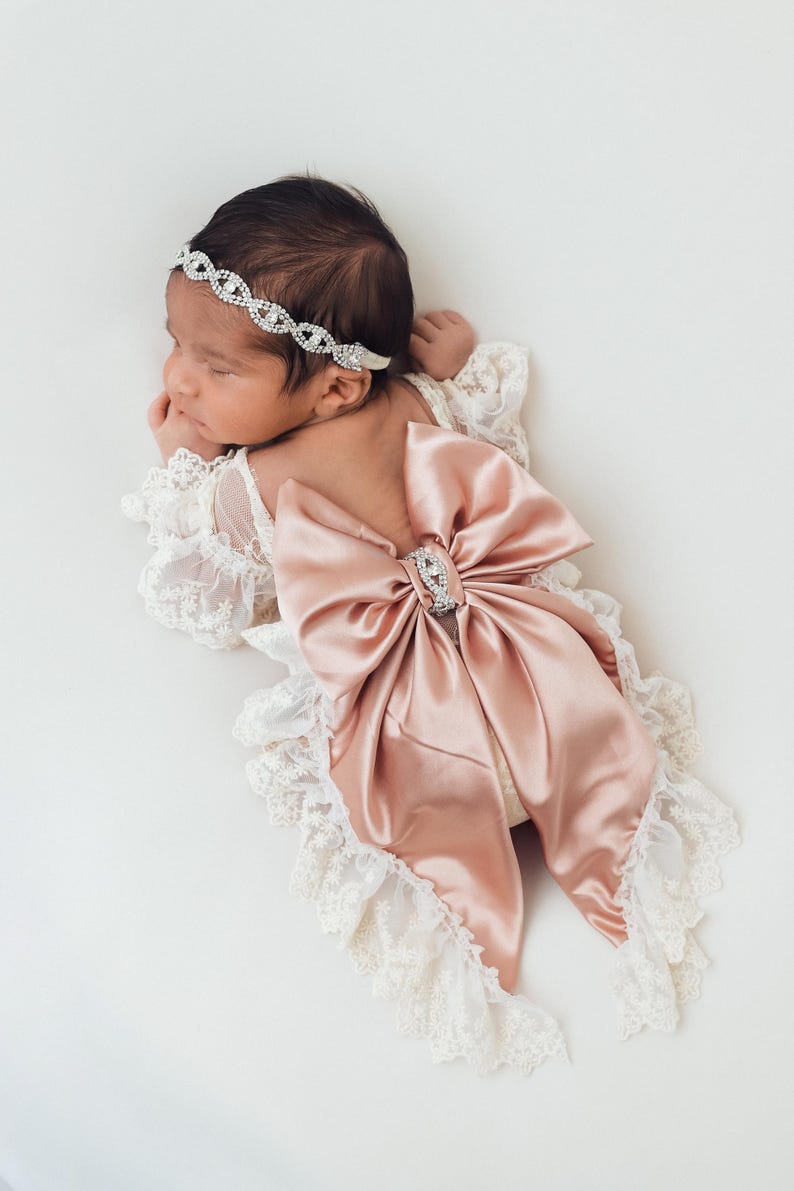 May include: A cream-colored lace romper featuring long sleeves with ruffled edges and a large, dusty rose satin bow on the back. The outfit is accented with a central rhinestone embellishment on the bow and a matching rhinestone headband.