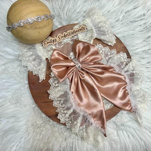 May include: A dusty rose satin bow featuring a central rhinestone embellishment, layered over white floral lace trim. A matching rhinestone headband rests on a wooden sphere. A wooden sign with the text Emilyz Embellishments is included.