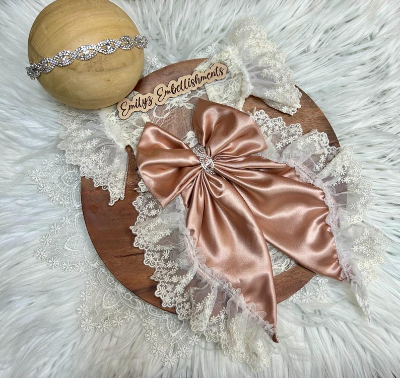 May include: A dusty rose satin bow featuring a central rhinestone embellishment, layered over white floral lace trim. A matching rhinestone headband rests on a wooden sphere. A wooden sign with the text Emilyz Embellishments is included.