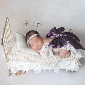 May include: A newborn infant wearing a white lace romper with a large dark purple satin bow on the back and a silver rhinestone headband, resting on a miniature cream-colored metal bed frame with a white pillow and a chunky knit cream blanket.