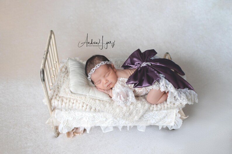 May include: A newborn infant wearing a white lace romper with a large dark purple satin bow on the back and a silver rhinestone headband, resting on a miniature cream-colored metal bed frame with a white pillow and a chunky knit cream blanket.