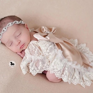 May include: A sleeping infant wearing a silver-toned rhinestone headband and a beige satin romper featuring intricate white lace overlays, ruffled lace trim along the edges, and a large satin bow detail on the back of the garment.