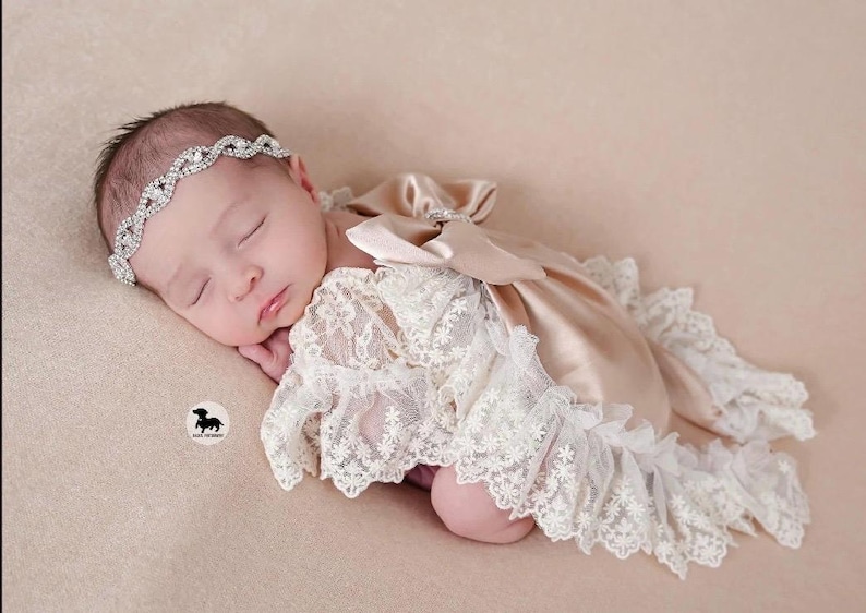 May include: A sleeping infant wearing a silver-toned rhinestone headband and a beige satin romper featuring intricate white lace overlays, ruffled lace trim along the edges, and a large satin bow detail on the back of the garment.