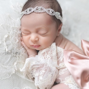May include: A sleeping infant wearing a silver-toned rhinestone headband and a white lace garment with a large, satin, dusty rose-colored bow attached to the side. The bow features a central rhinestone accent, and the lace has floral patterns.