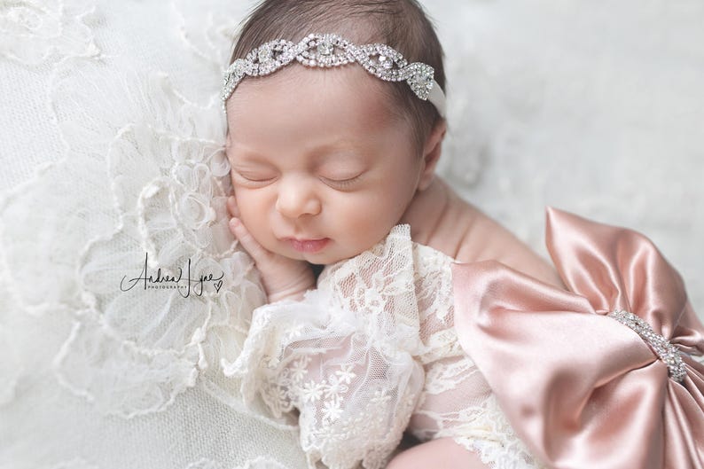 May include: A sleeping infant wearing a silver-toned rhinestone headband and a white lace garment with a large, satin, dusty rose-colored bow attached to the side. The bow features a central rhinestone accent, and the lace has floral patterns.