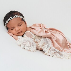 May include: A newborn infant wearing a cream-colored lace romper with long sleeves and a large, dusty rose satin bow on the back. The infant is also wearing a thin, silver-toned headband featuring a repeating circular rhinestone pattern.