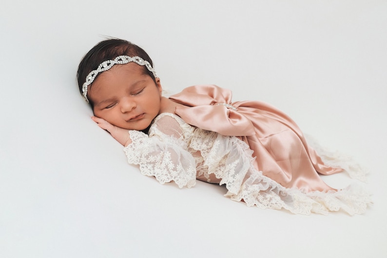 May include: A newborn infant wearing a cream-colored lace romper with long sleeves and a large, dusty rose satin bow on the back. The infant is also wearing a thin, silver-toned headband featuring a repeating circular rhinestone pattern.