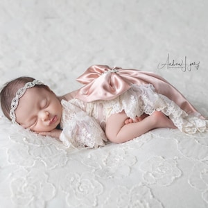 May include: A newborn infant wearing a cream-colored lace romper with a large, dusty pink satin bow on the back and a matching rhinestone-embellished headband. The infant is lying on a white lace fabric with a floral pattern.
