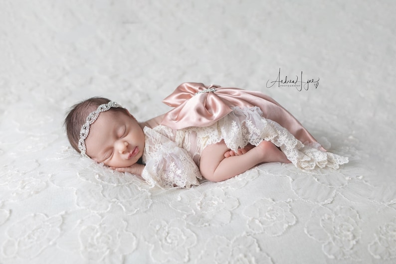 May include: A newborn infant wearing a cream-colored lace romper with a large, dusty pink satin bow on the back and a matching rhinestone-embellished headband. The infant is lying on a white lace fabric with a floral pattern.