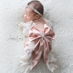 May include: A baby outfit featuring a large dusty rose satin bow with a central rhinestone embellishment, paired with cream-colored lace ruffles. The set includes a matching thin headband adorned with a series of interconnected rhinestone links.