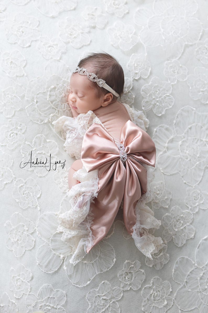 May include: A baby outfit featuring a large dusty rose satin bow with a central rhinestone embellishment, paired with cream-colored lace ruffles. The set includes a matching thin headband adorned with a series of interconnected rhinestone links.