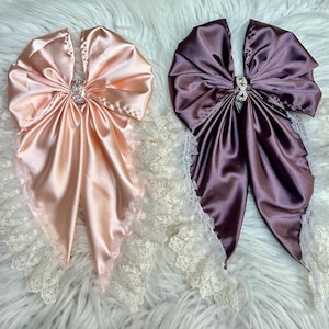 May include: Two large decorative bows made of smooth satin fabric, one in light peach and one in deep plum. Each bow features a central silver-toned rhinestone embellishment and is edged with a delicate white floral lace trim.
