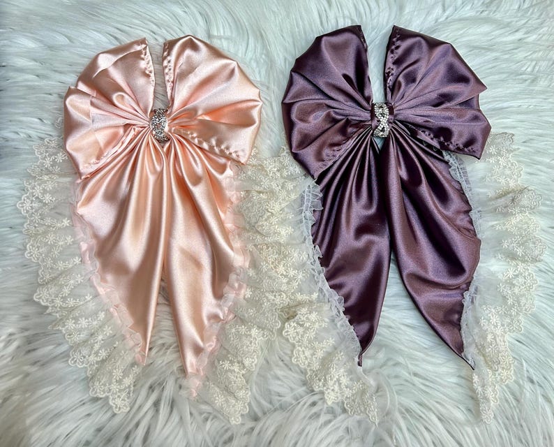 May include: Two large decorative bows made of smooth satin fabric, one in light peach and one in deep plum. Each bow features a central silver-toned rhinestone embellishment and is edged with a delicate white floral lace trim.