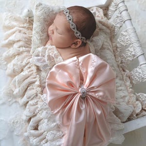 May include: A newborn infant wearing a pale pink satin bow with a central rhinestone embellishment and a matching rhinestone headband. The infant is resting on a cream-colored lace pillow inside a small white wooden bed frame with macrame details.