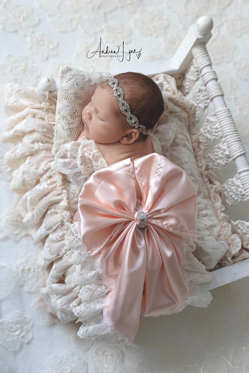 May include: A newborn infant wearing a pale pink satin bow with a central rhinestone embellishment and a matching rhinestone headband. The infant is resting on a cream-colored lace pillow inside a small white wooden bed frame with macrame details.