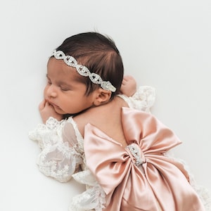 May include: A baby wearing a white lace long-sleeve garment with a large dusty pink satin bow on the back and a silver-toned rhinestone headband. The headband features an intricate crisscross pattern of clear crystals across the forehead.