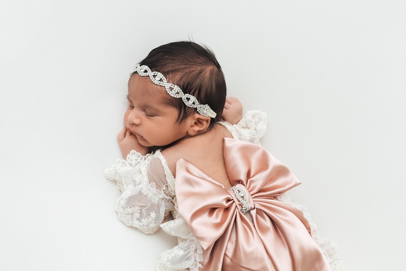 May include: A baby wearing a white lace long-sleeve garment with a large dusty pink satin bow on the back and a silver-toned rhinestone headband. The headband features an intricate crisscross pattern of clear crystals across the forehead.