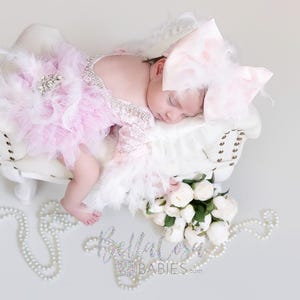 May include: A newborn infant wearing a light pink lace romper with white feather trim and a large matching pink bow headband. The infant is lying on a small white tufted bench next to a bouquet of white roses and strands of white faux pearls.
