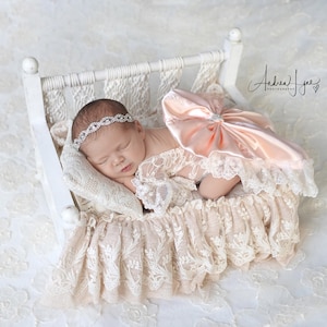 May include: A sleeping infant wearing a white lace outfit with a large peach-colored satin bow and a silver rhinestone headband, resting in a small white wooden bed frame draped with cream-colored lace fabric and a matching lace pillow.