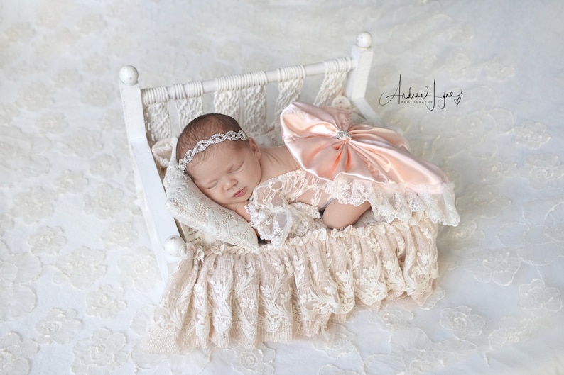 May include: A sleeping infant wearing a white lace outfit with a large peach-colored satin bow and a silver rhinestone headband, resting in a small white wooden bed frame draped with cream-colored lace fabric and a matching lace pillow.