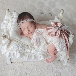 May include: A newborn infant sleeping on a small white bed with a ruffled white blanket and pillow. The infant wears a white lace outfit with a large satin dusty rose bow on the back and a silver-toned rhinestone headband across the forehead.