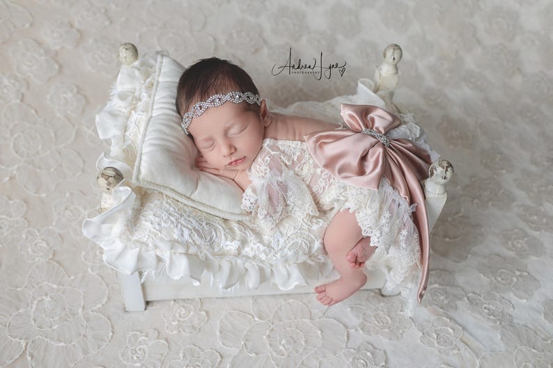 May include: A newborn infant sleeping on a small white bed with a ruffled white blanket and pillow. The infant wears a white lace outfit with a large satin dusty rose bow on the back and a silver-toned rhinestone headband across the forehead.