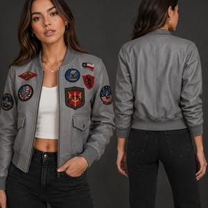 Puede incluir: Una chaqueta tipo bomber gris con cierre de cremallera frontal, puños y dobladillo acanalados. La parte delantera está decorada con varios parches bordados, incluyendo una bandera de Texas, un emblema circular de águila e insignias militares con texto.