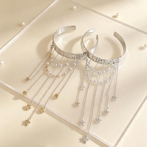 Peut inclure: Deux bracelets manchettes en métal argenté avec de multiples chaînes pendantes. L'un est orné de petites perles blanches et de breloques cœurs dorées, tandis que l'autre présente des étoiles argentées suspendues à de fines chaînes.