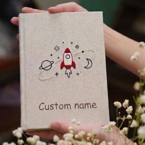 May include: A rectangular notebook with a beige fabric cover featuring an embroidered red and white rocket ship, a planet with rings, a crescent moon, and small stars. The text "Custom name" is embroidered in brown thread at the bottom center.