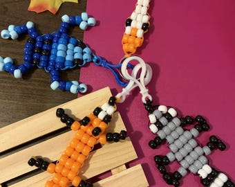 May include: Four lizard-shaped keychains crafted from plastic pony beads in various colour combinations: white and orange, blue and light blue, orange and black, and grey, black, and white. Each lizard features a plastic clip for attachment.