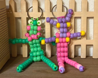 May include: Two handmade keychains crafted from colourful plastic pony beads. The left keychain is primarily green with pink accents and light-coloured eyes. The right keychain features a purple and yellow head with a pink body and purple limbs.