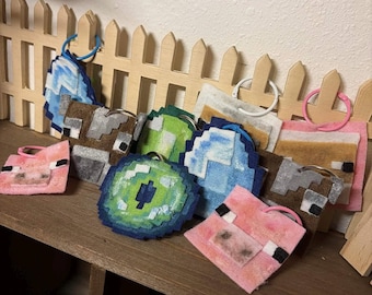May include: A collection of square and circular felt keychains featuring pixelated designs in various colours including pink, brown, green, and blue. Each piece is attached to a colourful plastic ring, showcasing blocky patterns reminiscent of digital game graphics.