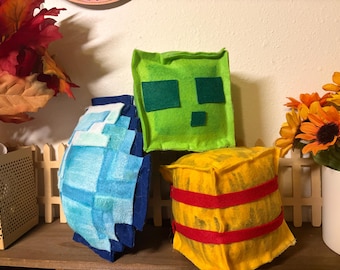 May include: Three plush toys made of felt. One is a light blue and dark blue pixelated shape. Another is a square lime green block with dark green felt eyes and a mouth. The third is a yellow cube with two horizontal red felt stripes wrapped around it.