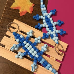 May include: Two lizard-shaped keychains crafted from light blue and dark blue plastic pony beads. Each lizard features a long tail, four legs, and a metal split ring attached to the head or tail for use as a functional keychain accessory.