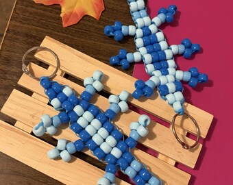May include: Two lizard-shaped keychains crafted from light blue and dark blue plastic pony beads. Each lizard features a long tail, four legs, and a metal split ring attached to the head or tail for use as a functional keychain accessory.