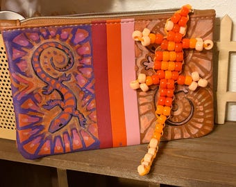May include: A rectangular zippered pouch featuring a purple and orange lizard illustration on the left, vertical stripes in shades of orange and pink in the centre, and a brown section on the right with a three-dimensional orange beaded lizard.