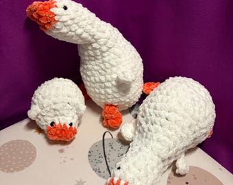 May include: Three handmade crochet plush toys shaped like geese and a gosling. The figures are crafted from white, textured yarn with bright orange beaks and feet. Each plush features small black bead eyes and a soft, rounded, stuffed body.