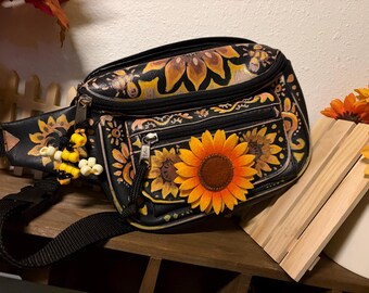 May include: A black bum bag featuring hand-painted yellow and orange sunflower motifs, a front zippered pocket, and a large fabric sunflower attached to the front. A small beaded bee charm hangs from the main zipper pull, and the bag has a black strap.
