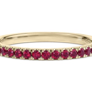 May include: A thin gold band ring featuring a continuous row of small, round, deep red rubies set along the front half of the metal surface. The band has a polished finish, and the gemstones are held in place by small, uniform metal prongs.