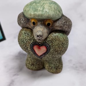 May include: A ceramic figurine of a stylized poodle with a textured green and gray coat. The poodle has large yellow eyes, a light-colored snout, and a prominent heart-shaped emblem on its chest featuring a red center and a dark decorative border.