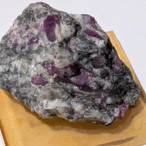 May include: A raw mineral specimen featuring deep magenta-colored crystals embedded within a matrix of grey, black, and white stone. The rock has a rough, uneven texture with visible crystalline structures throughout the dark and light mineral layers.