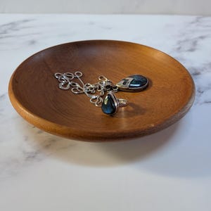 May include: A round, smooth, medium-brown wooden bowl containing a silver necklace with a teardrop-shaped dark stone pendant and a matching silver ring featuring a teardrop-shaped dark stone, both displaying iridescent blue flashes.