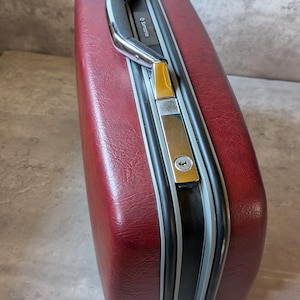 Pode incluir: Uma mala de viagem rígida Samsonite vintage na cor vermelho bordô profundo com acabamento texturizado. A bagagem apresenta uma alça de plástico preta, ferragens de metal prateado, um mecanismo de fecho central com fechadura e detalhes em preto.