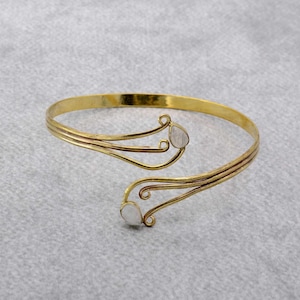 May include: A gold-toned open cuff bracelet featuring a polished metal band that splits into two curved, wire-like ends. Each terminal is accented with a small, smooth, teardrop-shaped white stone, creating a symmetrical and open-ended design.