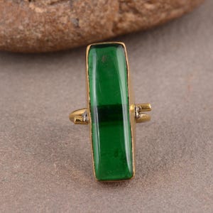 May include: A gold-toned ring featuring a long, rectangular, translucent green stone set vertically. The metal band is thin and connects to the sides of the stone setting, which has a smooth, polished finish and a vibrant, deep green color.
