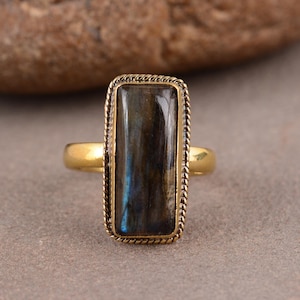 May include: A gold-toned ring featuring a rectangular labradorite gemstone with blue and dark gray tones. The stone is set in a twisted rope-style metal bezel, mounted on a smooth, polished gold-toned band that encircles the finger.