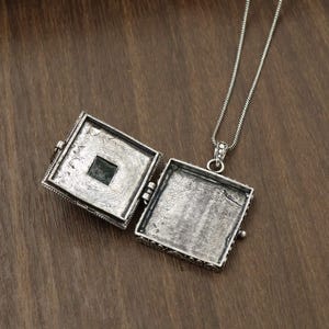 May include: A square silver locket pendant with an oxidized finish, shown in an open position to reveal two empty interior compartments. The locket is attached to a thin silver chain, featuring a textured border and a small dark square inlay.
