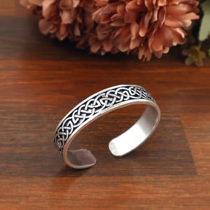May include: A silver-toned cuff bracelet featuring an intricate, repeating Celtic knot pattern engraved along the outer surface. The metal band has an open design with smooth, rounded ends, showcasing a polished finish with darkened recessed details.